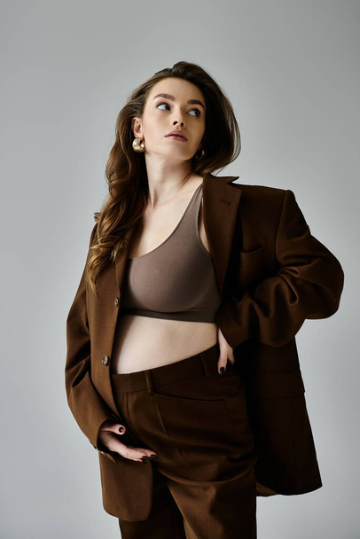 Une jeune femme enceinte en costume marron avec un blazer frappant une pose confiante sur un fond gris. - Photo, image