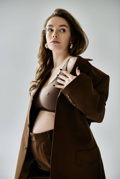 Стильная молодая беременная женщина в коричневом костюме в блейзере позирует на сером фоне. - Фото, изображение