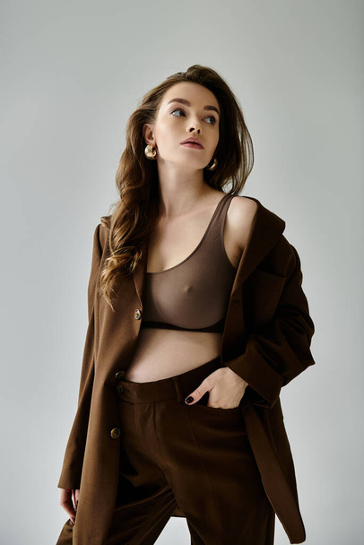 Una joven embarazada emana confianza en un traje marrón de moda con una chaqueta, posando sobre un fondo gris. - Foto, imagen