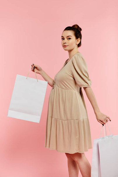 Eine junge schwangere Frau in einem Kleid hält freudig zwei Einkaufstüten vor rosa Hintergrund. - Foto, Bild