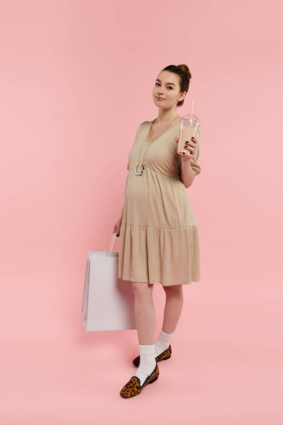 Młoda ciężarna kobieta w pięknej sukience trzymająca drinka i torby na zakupy na różowym tle. - Zdjęcie, obraz