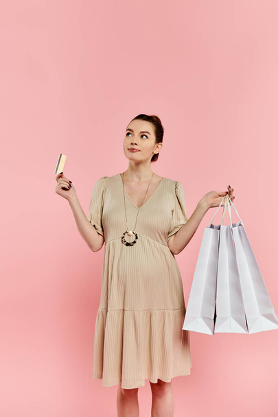 Una donna incinta elegante in un vestito che bilancia due borse della spesa e una carta di credito su uno sfondo rosa. - Foto, immagini