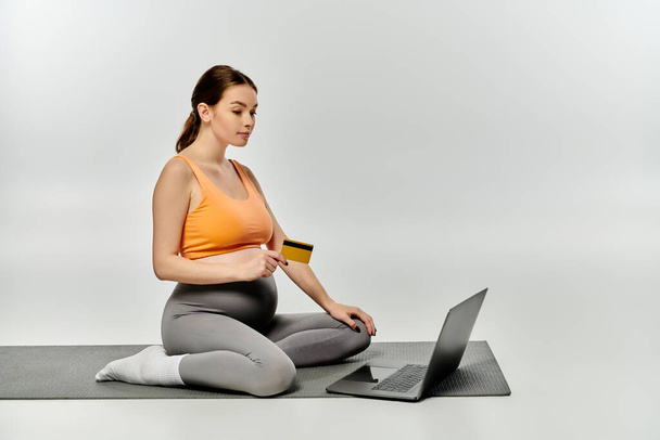 Egy terhes nő aktív ruhában ül egy jóga szőnyegen, miközben egy laptopon dolgozik.. - Fotó, kép