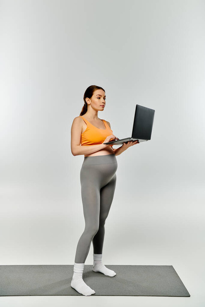 Sportowa ciężarna kobieta stoi na macie, trzymając laptopa z ostrością i determinacją. - Zdjęcie, obraz