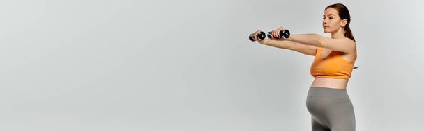 A pregnant woman in activewear confidently lifts dumbbells in front of a white background. - Photo, Image