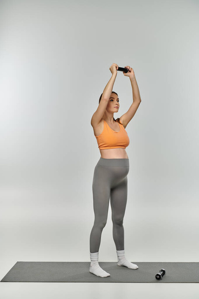 Een zwangere vrouw in activewear staat op een yogamat met opgeheven handen in de lucht en straalt sereniteit en gratie uit. - Foto, afbeelding