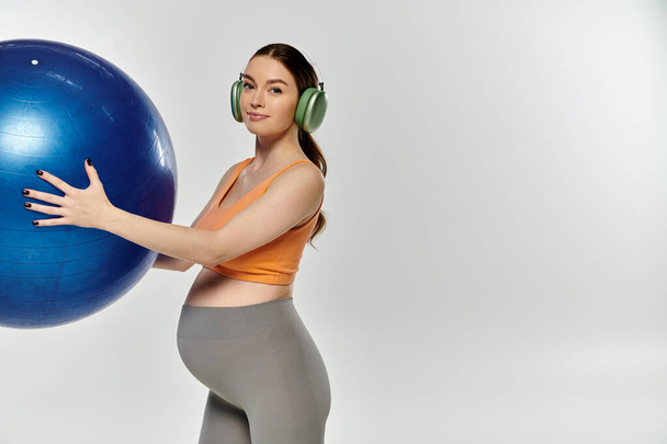 Una mujer embarazada y deportiva en uso activo equilibra una gran bola azul en una mano mientras sostiene los auriculares en la otra. - Foto, imagen