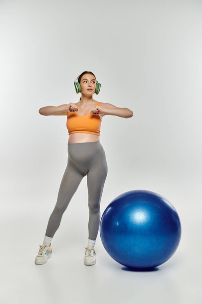 Беременная женщина в спортивной одежде стоит рядом с ярко-синим мячом для упражнений на сером фоне. - Фото, изображение