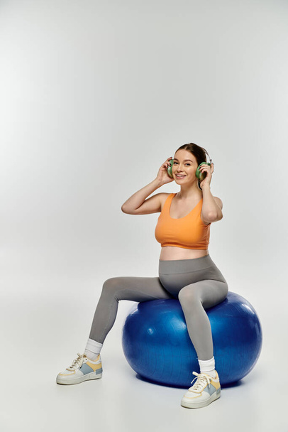 Young pregnant woman in active wear balances on blue ball, deeply engaged in music. - Photo, Image