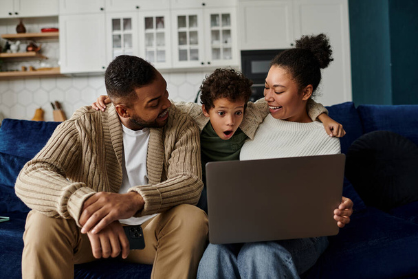 Un homme et une femme sont assis sur un canapé avec un enfant, tous regardant un ordinateur portable. - Photo, image