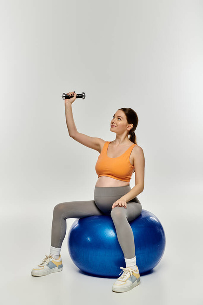 Egy aktív ruhás terhes nő egyensúlyt mutat azzal, hogy egy labdán ül, miközben egy súlyzót tart a kezében.. - Fotó, kép