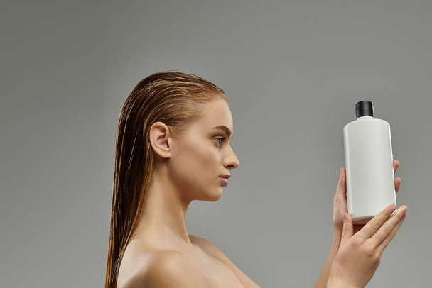 Молодая женщина с мокрыми волосами нежно держит в руке бутылку шампуня.. - Фото, изображение