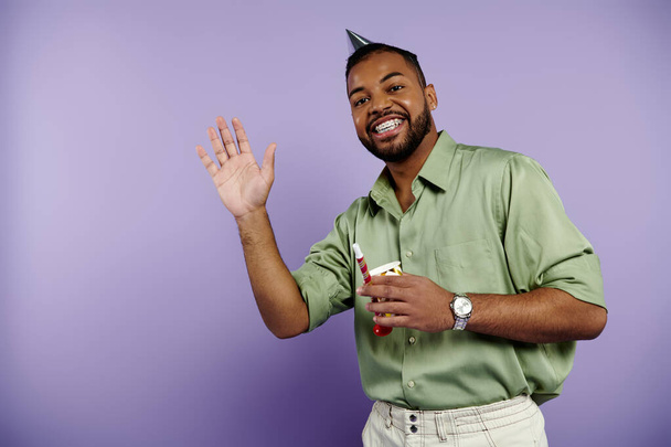Young African American man with braces happily raises his hand on a purple background. - Photo, Image