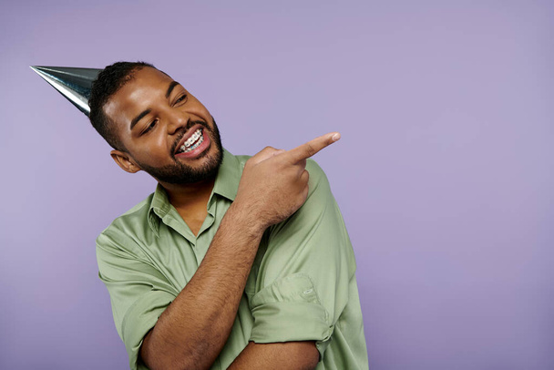 Ein junger afroamerikanischer Mann mit Hosenträgern zeigt begeistert, während er einen festlichen Partyhut vor lila Hintergrund trägt. - Foto, Bild