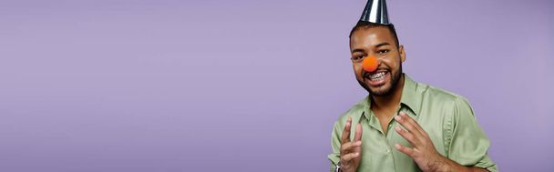 紫色のバックグラウンドに勇敢な若いアフリカ系アメリカ人男性は幸せに彼の頭の上にピエロの鼻のバランスをとっています. - 写真・画像