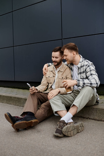 Двое мужчин в повседневной одежде сидят на земле, поглощенные сотовыми телефонами. - Фото, изображение
