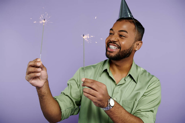 Un joven afroamericano con frenos sosteniendo alegremente chispas mientras usa un sombrero de fiesta sobre un fondo púrpura. - Foto, Imagen