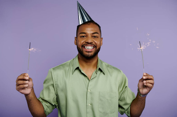 若い黒人男性は笑顔で,紫色の背景にパーティー帽子をかぶった2つのスパークラーを保持. - 写真・画像