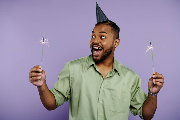Junger afroamerikanischer Mann mit Zahnspange hält lächelnd zwei Wunderkerzen im Partyhut vor leuchtend violettem Hintergrund. - Foto, Bild