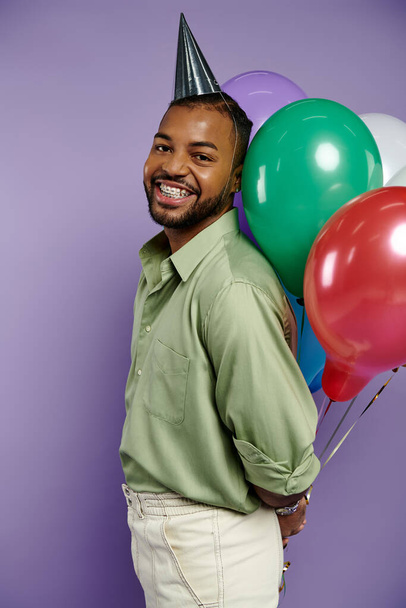 Young African American man in a party hat happily holds colorful balloons on a purple background. - Фото, изображение