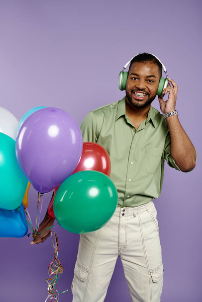Jonge Afro-Amerikaanse man met beugel glimlacht, houdt ballonnen en koptelefoon tegen een paarse achtergrond. - Foto, afbeelding