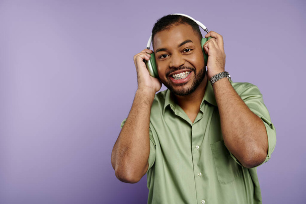 Un joven afroamericano feliz con frenos sonríe ampliamente para la cámara mientras usa auriculares sobre un vibrante fondo púrpura. - Foto, imagen