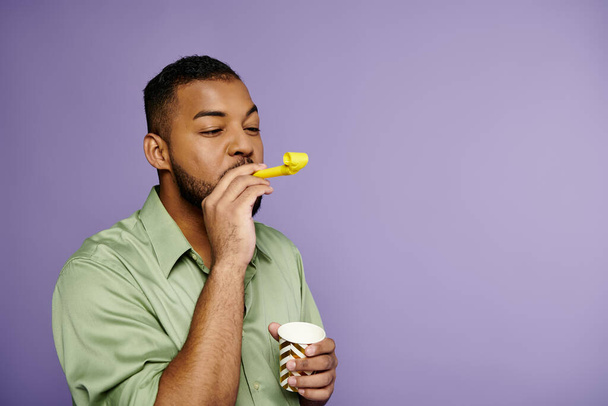 Un jeune Afro-Américain dans une chemise verte soufflant corne de fête sur un fond violet. - Photo, image