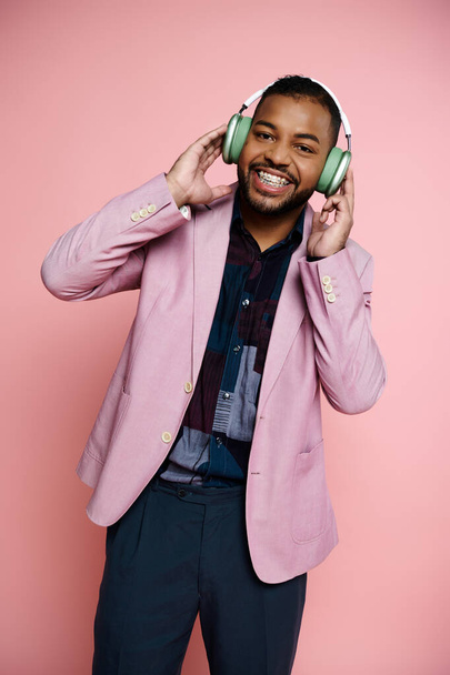 Un joven y feliz afroamericano con aparatos ortopédicos escucha auriculares con una chaqueta rosa sobre un fondo rosa. - Foto, imagen