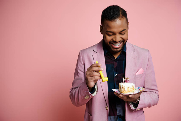 Egy elegáns fiatal afro-amerikai férfi rózsaszín öltönyben boldogan tart egy tányér szülinapi tortát és party kürtöt élénk rózsaszín háttér mellett.. - Fotó, kép