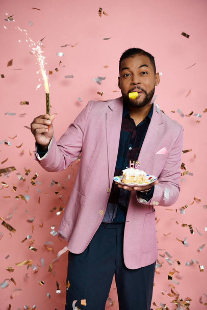 Joven hombre afroamericano con chaqueta rosa sostiene alegremente un pastel y chispeante en un momento de celebración. - Foto, imagen