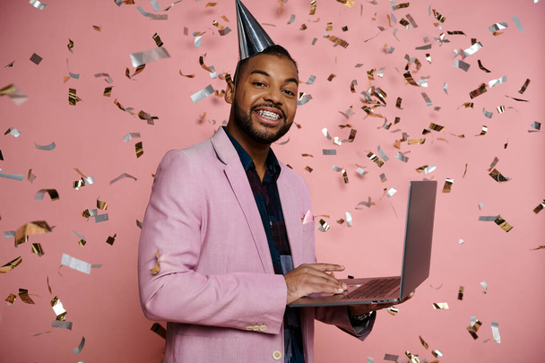 Joyful young African American man in braces balancing a laptop, dressed in a party hat on a vibrant pink background. - Foto, immagini