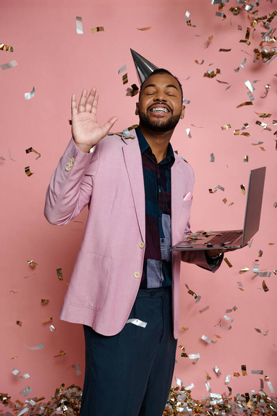 Een jonge Afro-Amerikaanse man met een beugel glimlacht terwijl hij een laptop vasthoudt en een feestmuts op een roze achtergrond draagt. - Foto, afbeelding