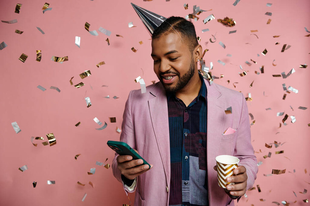 Młody Afroamerykanin w aparacie na zęby, szczęśliwy, trzymający kubek i telefon komórkowy, stojący w kapeluszu imprezowym na różowym tle. - Zdjęcie, obraz