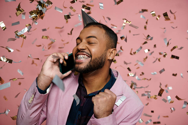 Junger afroamerikanischer Mann in Hosenträgern trägt einen Partyhut und spricht fröhlich auf einem Handy vor rosa Hintergrund. - Foto, Bild