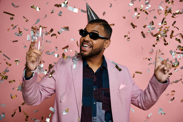 A young African American man in braces, looking happy, wears a party hat and sunglasses on a pink background. - Photo, Image