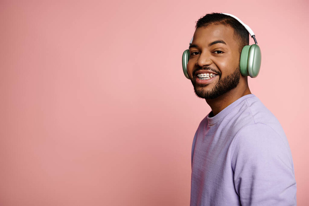 Joven hombre afroamericano con frenos sonriendo brillantemente mientras usa auriculares sobre un fondo rosa. - Foto, imagen