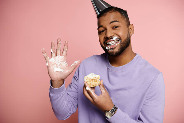 Young African American man in party hat happily eating a birthday cake on a pink background. - Photo, Image