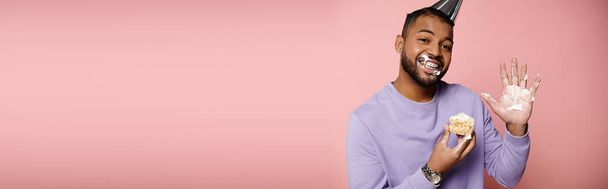 Щасливий, молодий афроамериканський чоловік з брекетами, у фіолетовій сорочці, смакує випічку на рожевому фоні. - Фото, зображення