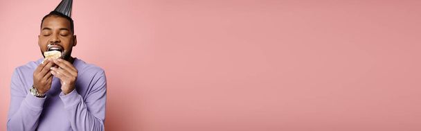 Молодой афроамериканец с брекетами радостно ест праздничный торт в шляпе на розовом фоне.. - Фото, изображение