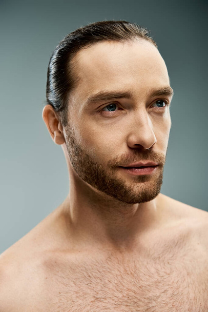 Un hombre sin camisa que emana masculinidad y confianza, mostrando su barba rugosa en un estudio sobre un fondo gris. - Foto, Imagen