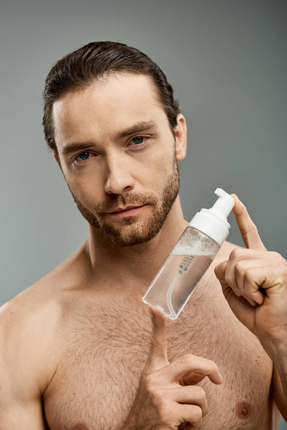 Un hombre guapo y sin camisa con barba sostiene con gracia una botella de limpiador contra un fondo gris del estudio. - Foto, imagen