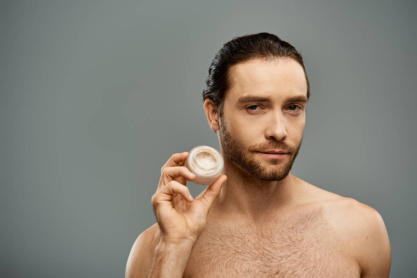 Uomo senza maglietta con la barba che tiene un barattolo di crema su uno sfondo grigio in un ambiente da studio. - Foto, immagini