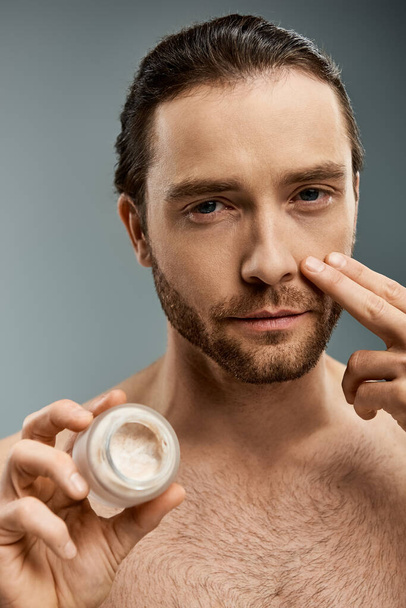 Handsome shirtless man with a beard holding a jar of cream against a grey backdrop. - Photo, Image