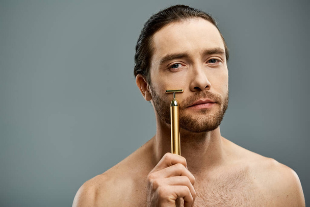 Homme torse nu avec barbe tenant un rasoir doré devant le visage sur fond gris. - Photo, image