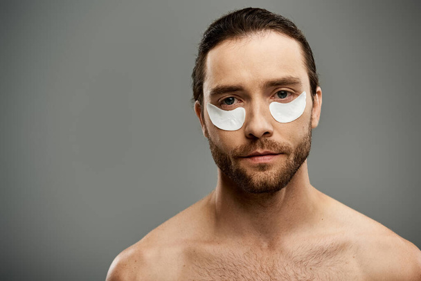 Um homem bonito e sem camisa com uma barba confiantemente usa manchas nos olhos em uma pose impressionante em um fundo de estúdio cinza. - Foto, Imagem