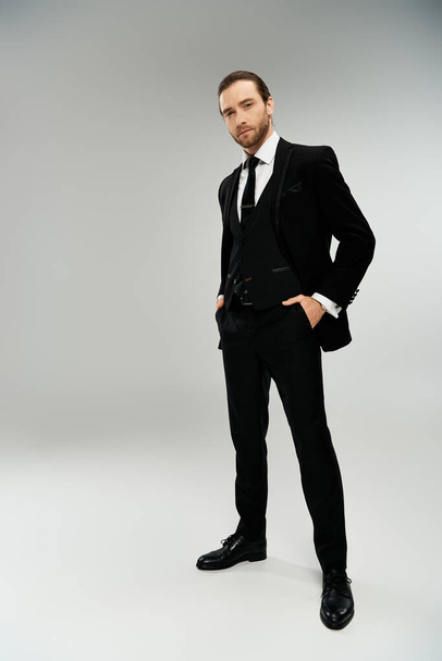 Un hombre de negocios barbudo con un esmoquin elegante posa con confianza sobre un fondo gris de estudio. - Foto, imagen