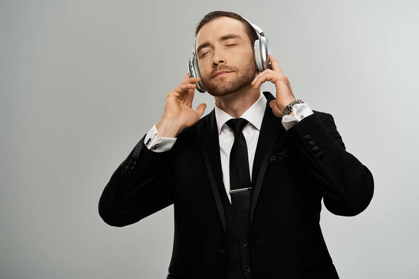Een bebaarde zakenman in een pak luistert aandachtig naar een koptelefoon tegen een grijze studio achtergrond. - Foto, afbeelding