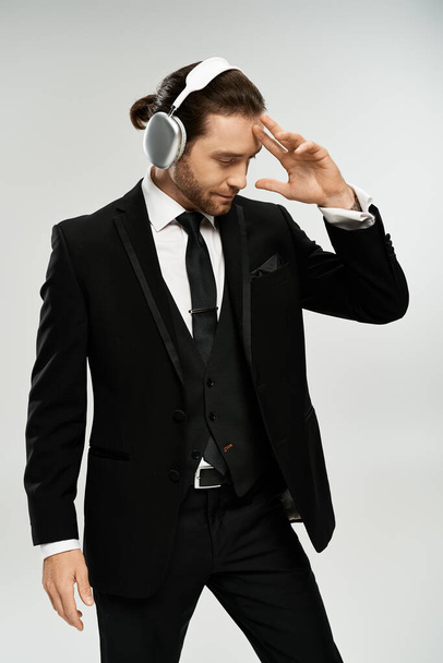 Een bebaarde, knappe zakenman in een pak die naar muziek luistert met een koptelefoon in een studio op een grijze achtergrond. - Foto, afbeelding