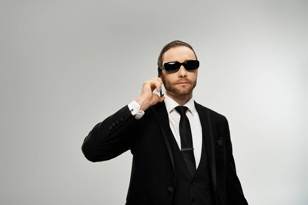 Vousatý podnikatel v obleku a slunečních brýlích vede telefonickou konverzaci na pozadí šedého studia. - Fotografie, Obrázek
