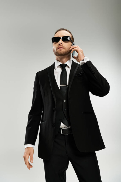 Γενειοφόρος επιχειρηματίας με κομψό κοστούμι και γυαλιά ηλίου συνομιλεί σε ένα κινητό τηλέφωνο, αποπνέοντας εμπιστοσύνη και επαγγελματισμό. - Φωτογραφία, εικόνα